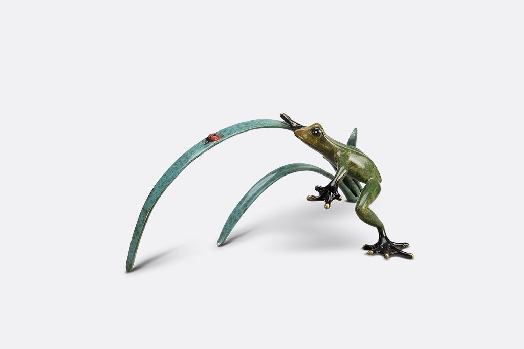 A green frog figurine on top of long blue-green leaves ready to pounce a lady bug.