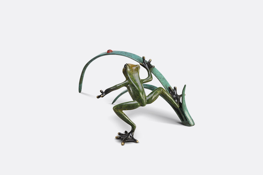 A green frog figurine on top of long blue-green leaves ready to pounce a lady bug.