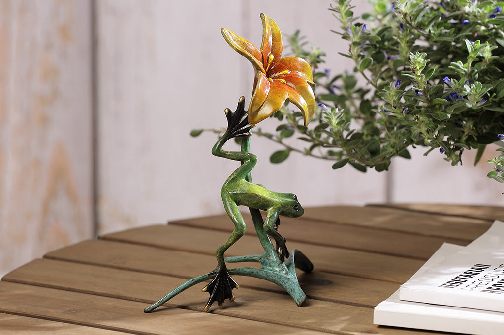 The side of a statue of a frog climbing around a yellow flower placed on a table.