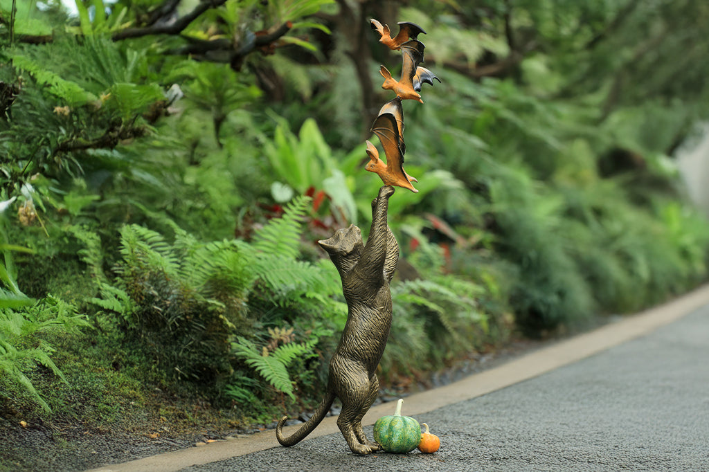cast metal cat sculpture on back paw reaching/ playing with orange and black bats with two pumpkins by base of kitty hind paws. Set in garden scene
