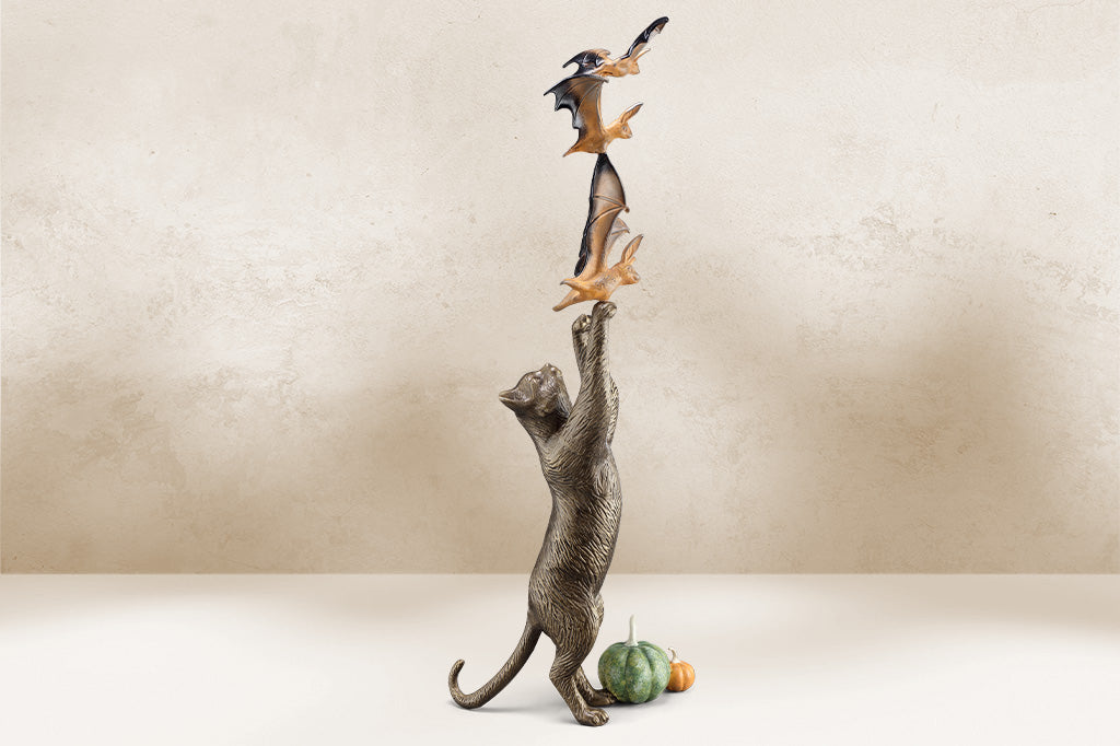cast metal cat sculpture on back paw reaching/ playing with orange and black bats with two pumpkins by base of kitty hind paws