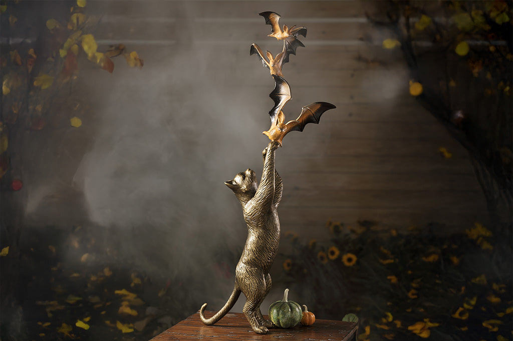 orange and black bats in flight above a bronze cat sculpture, standing on hind legs with two cast metal pumpkins at its feet. shown in foggy, autumn outdoor scene