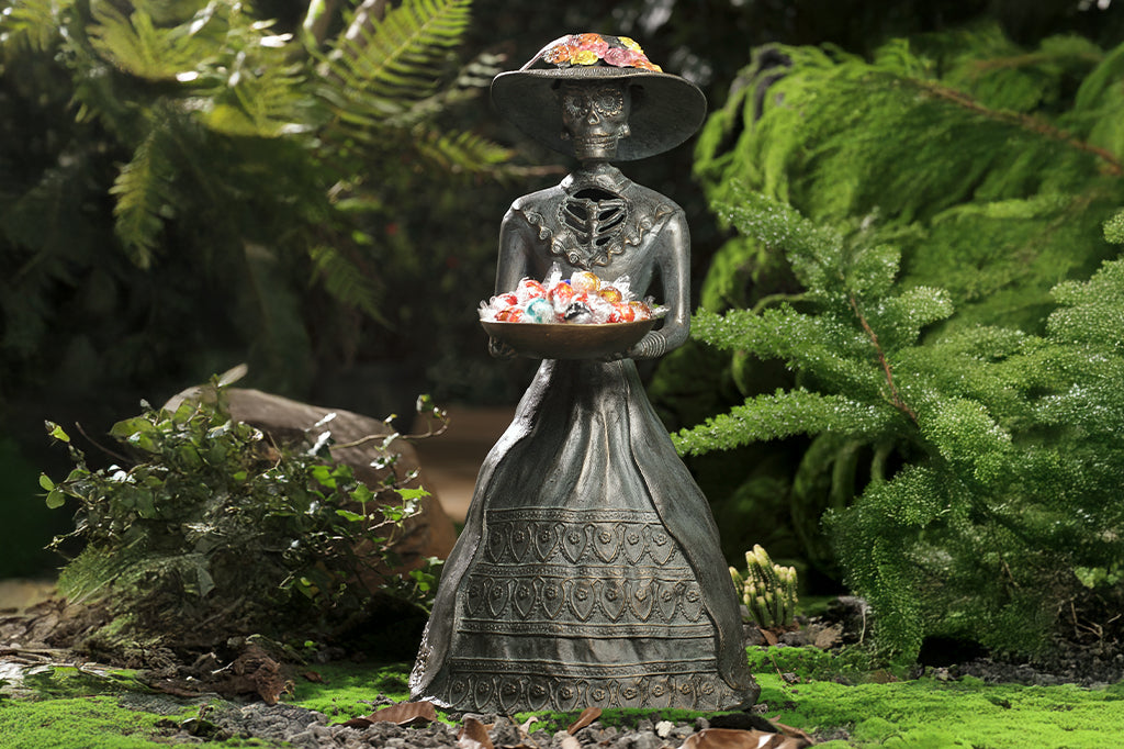 cast metal sculpture of la catrina with decorative bowl holding candies in garden setting