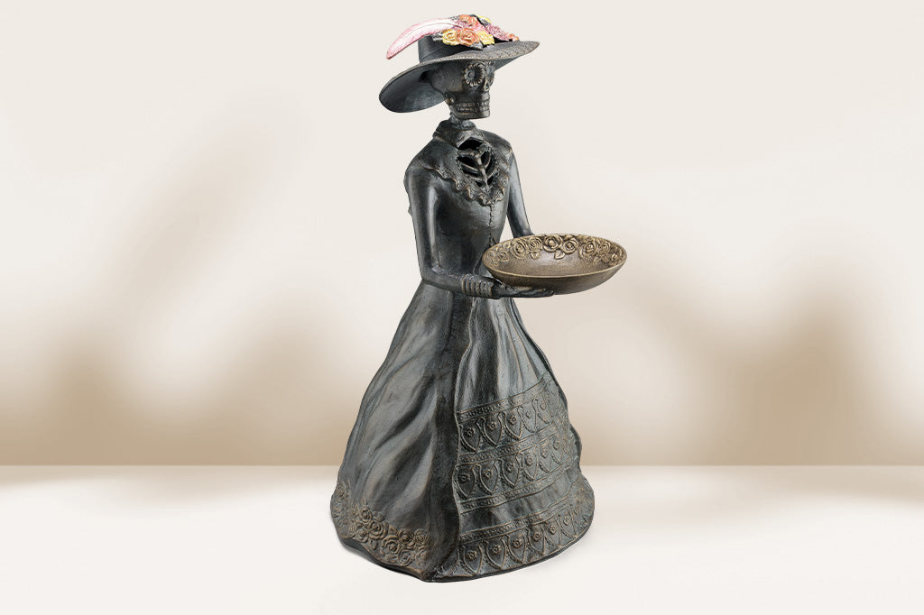cast metal sculpture of la catrina skeleton in dress and hat with decorative bowl