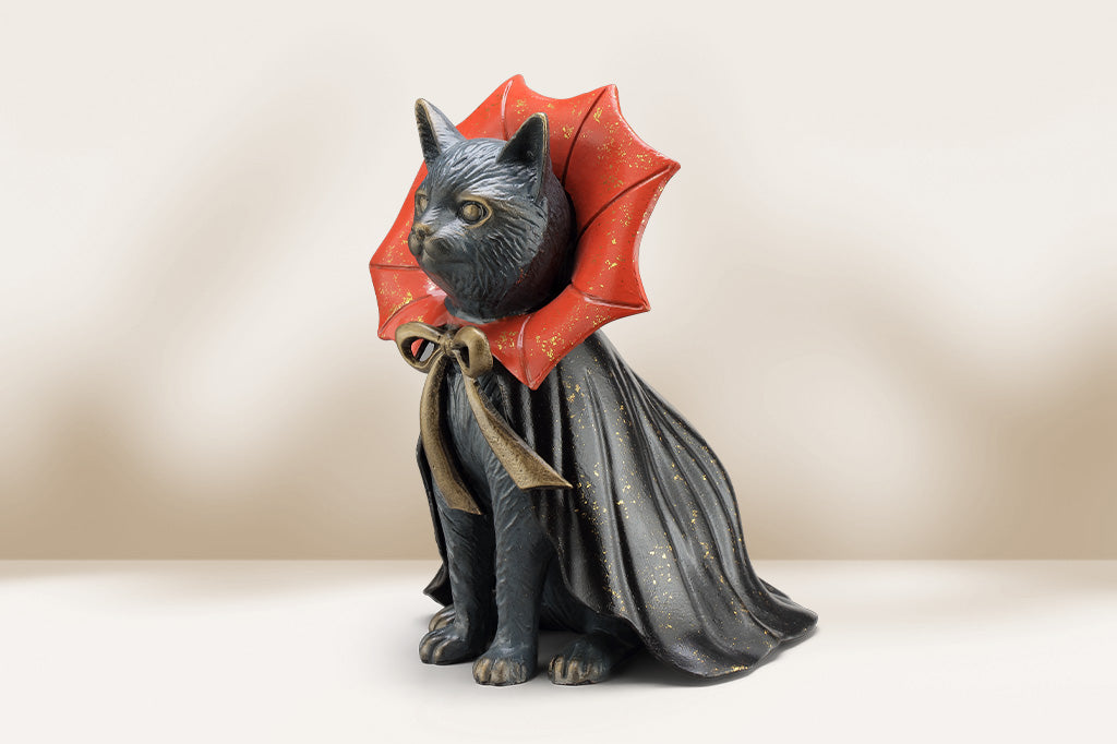Cat sculpture of cast metal wearing a red and black vampire robe on a beige background. flared collar is shaped like bat wings and tied with bronze bow.