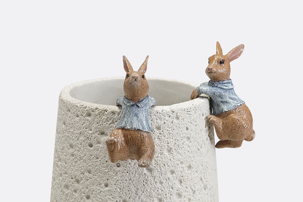 A hanging sculpture of two bunnies wearing blue shirts that dangle off of plant pots.