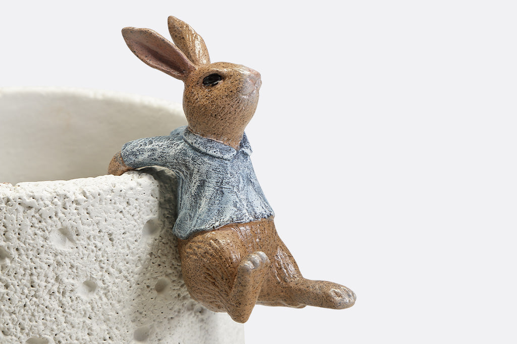 A hanging sculpture of two bunnies wearing blue shirts that dangle off of plant pots.