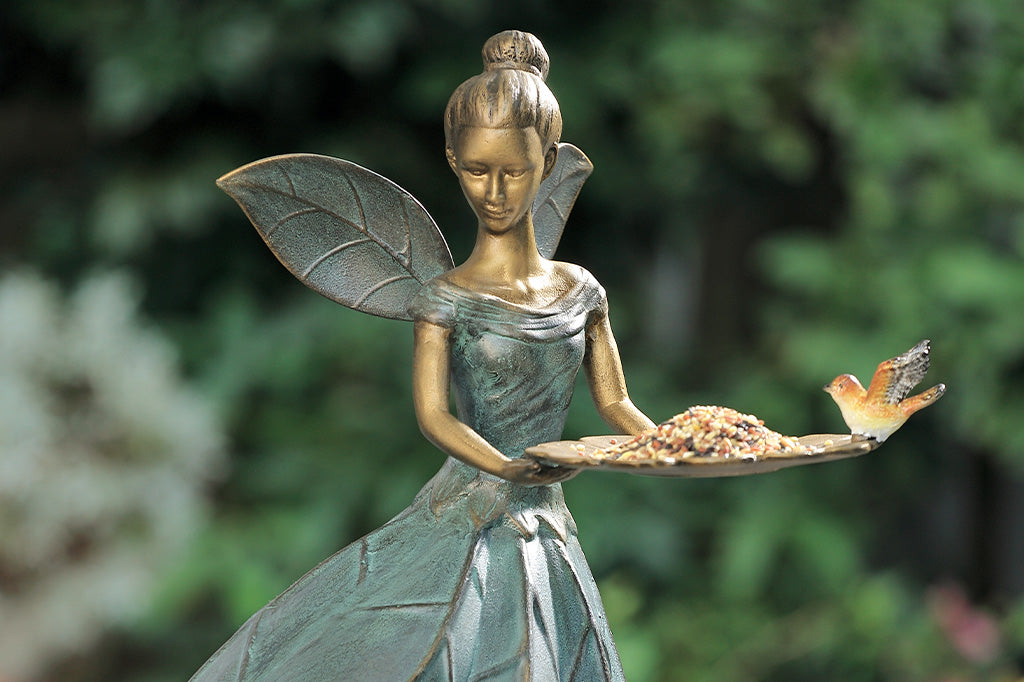 garden fairy statue birdfeeder holding a tray with a bird and flowers against a blurred green background. tray holds birdseed
