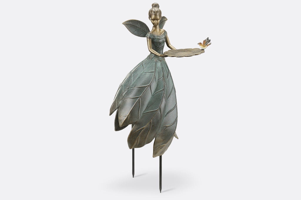 Cast metal garden statue of a fairy with leaf-like wings and dress, held up by removable stakes. shown on white background. Holds a bird feeder shaped like blossom and adorned with bird