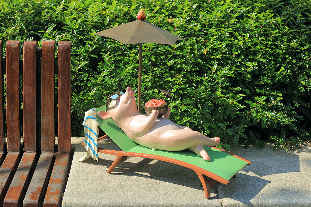 Pig sculpture sunbathing on a lounge chair with an umbrella, set against a green hedge.