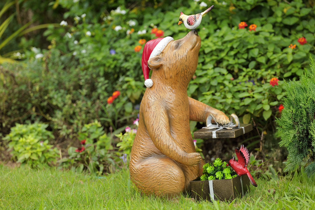 Bear statue with a bird on its head, sitting in front of holder for plants with a gift box shape, in a garden setting.