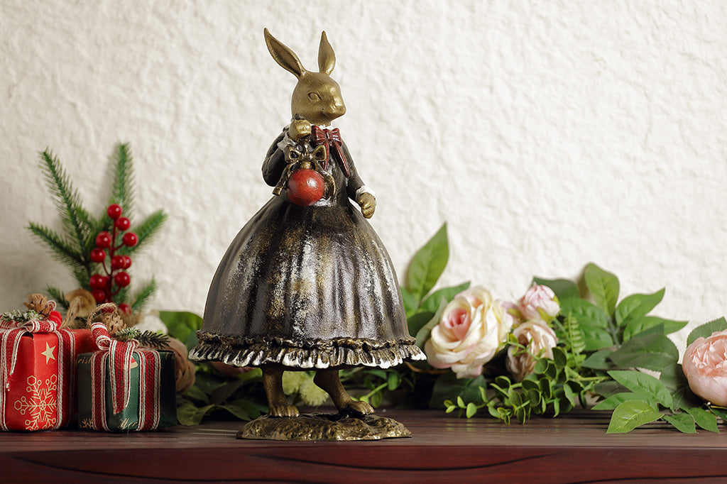 Sculpture of a bunny dressed in Victorian Era attire holding a red Chirstmas ornament.
