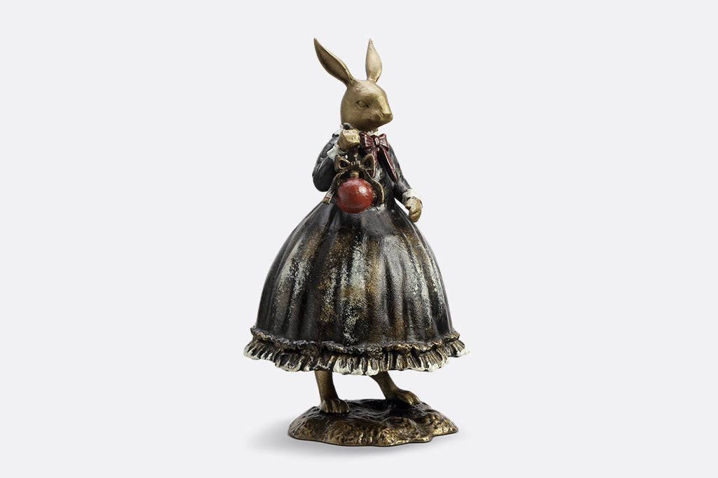 Sculpture of a bunny dressed in Victorian Era attire holding a red Chirstmas ornament.