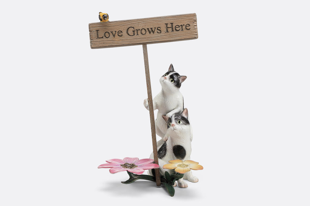 A garden sculpture of two cats holding a sign that says "love grows here" with a bird sitting on the sign and a pair of flowers at the bottom.