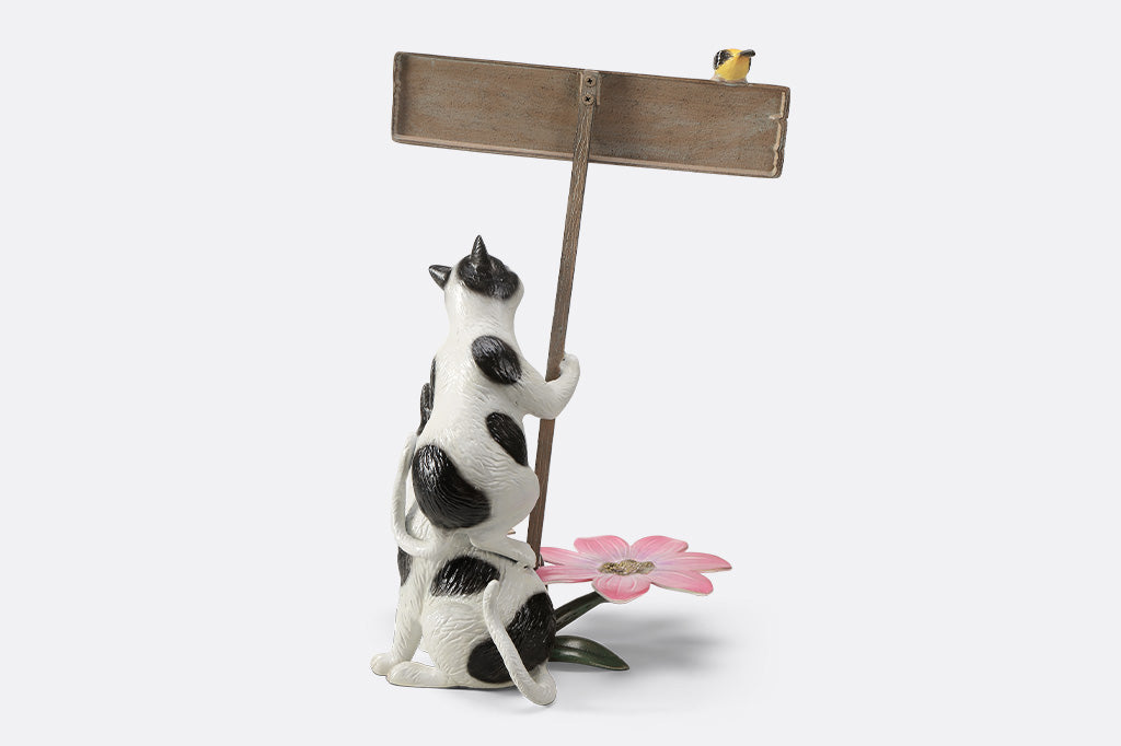 A garden sculpture of two cats holding a sign that says "love grows here" with a bird sitting on the sign and a pair of flowers at the bottom.