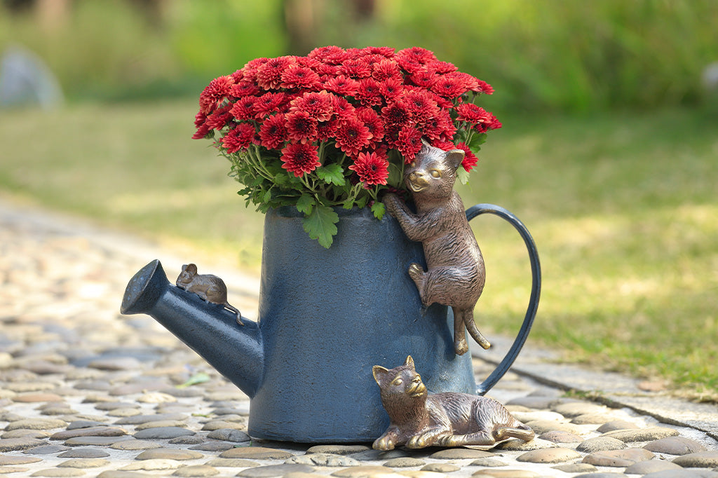 A decorative watering can planter with two cats climbing around to follow a mouse.
