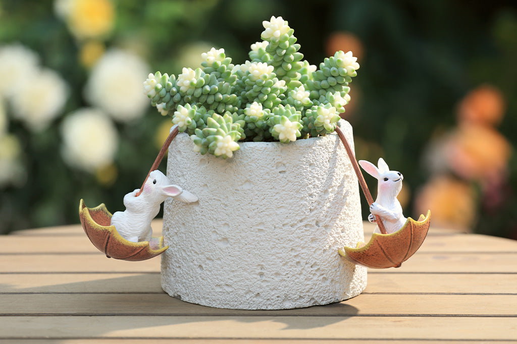 Pot hanger sculptures of two white bunnies in amber colored umbrellas. One bunny is facing left and the other is facing straight ahead.