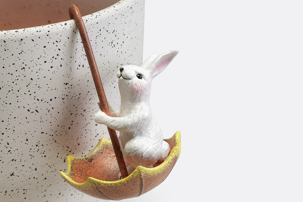 Pot hanger sculptures of two white bunnies in amber colored umbrellas. One bunny is facing left and the other is facing straight ahead.