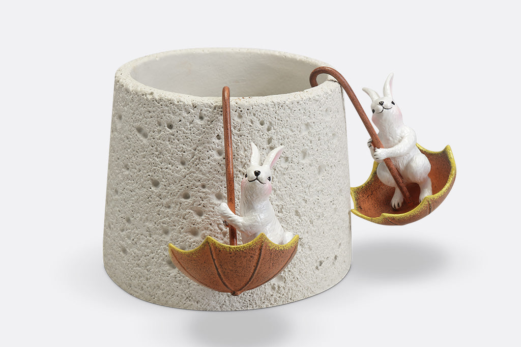 Pot hanger sculptures of two white bunnies in amber colored umbrellas. One bunny is facing left and the other is facing straight ahead.