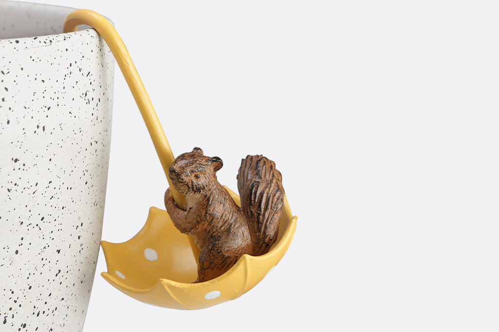 A hanging pot sculpture of a squirrel playing with a yellow umbrella.