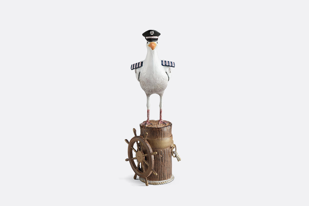 A sculpture of a seagull dressed in Captain regalia standing atop a dock adorned with rope and a ship's steering wheel.