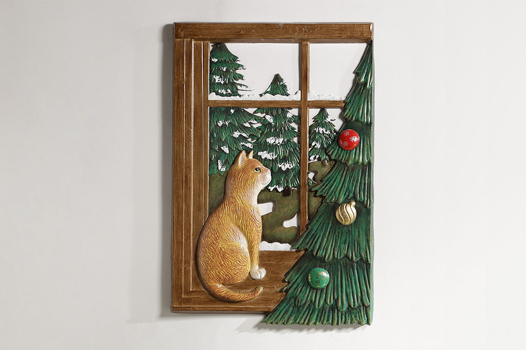 A decorative wall art of a cat looking out a window at trees next to a Chirstmas tree.