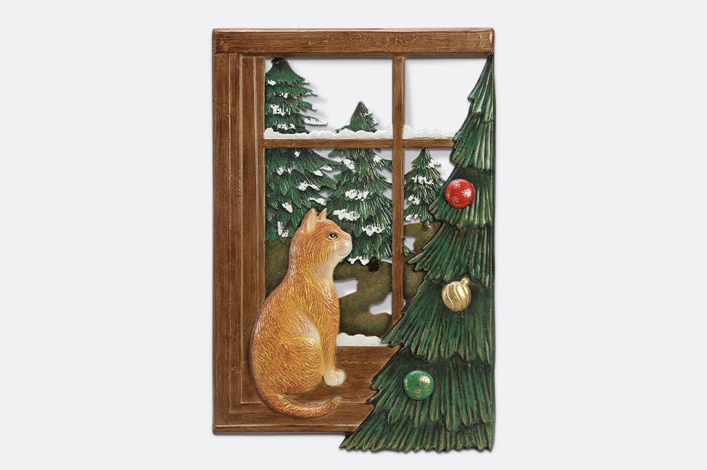 A decorative wall art of a cat looking out a window at trees next to a Chirstmas tree.