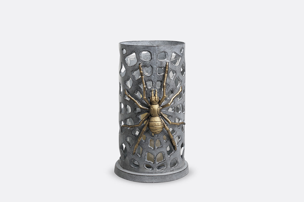 A metallic spider figurine mounted on a perforated metal cylinder holder, set against a plain light gray background.