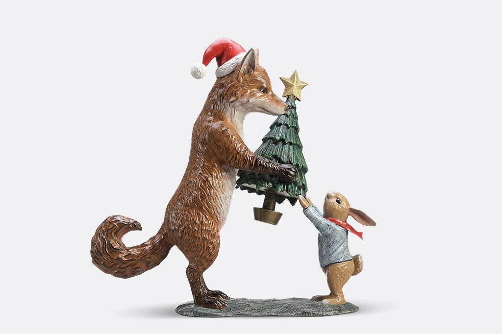 A sculpture of a fox wearing a santa hat is giving a rabbit with a scarf a christmas tree with a star on top.