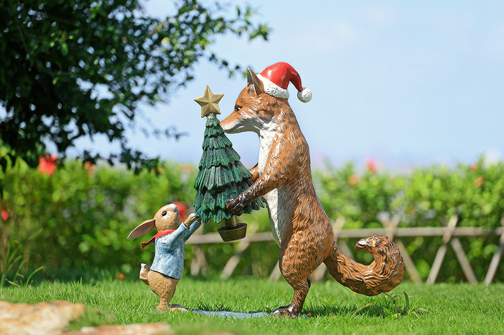 A sculpture of a fox wearing a santa hat is giving a rabbit with a scarf a christmas tree with a star on top.