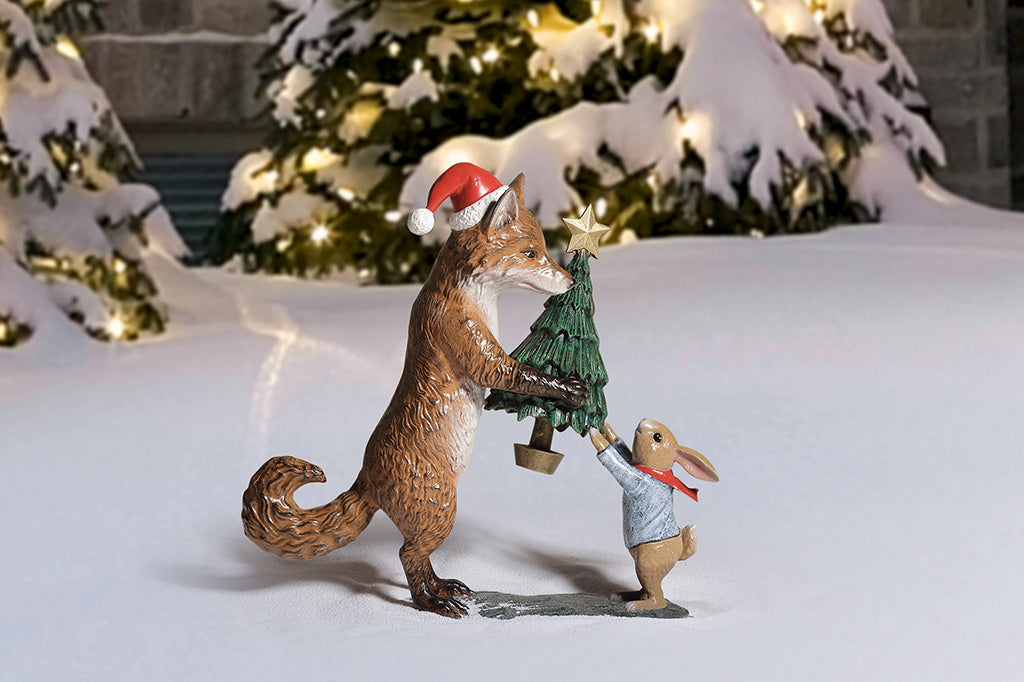 A sculpture of a fox wearing a santa hat is giving a rabbit with a scarf a christmas tree with a star on top.