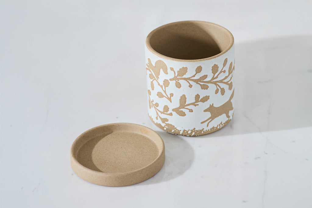 A white cylindrical container with a matching round lid, decorated with a tan botanical and deer pattern, set on a light gray marble surface.