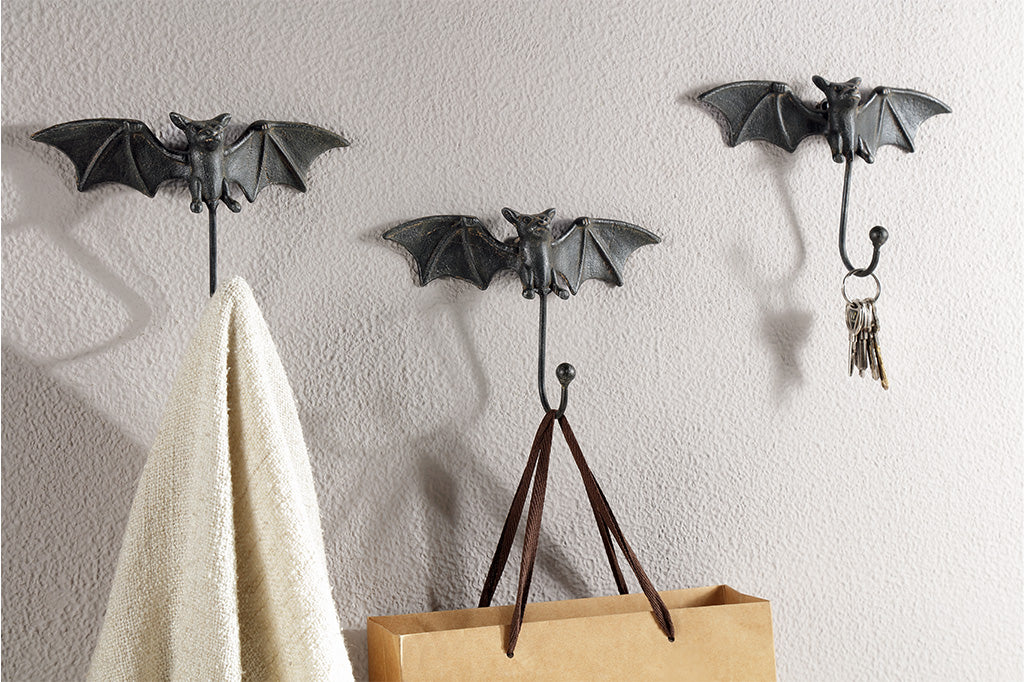 3 bat shaped wall hooks of cast iron shown on wall holding a scarf, tote bag, and keys