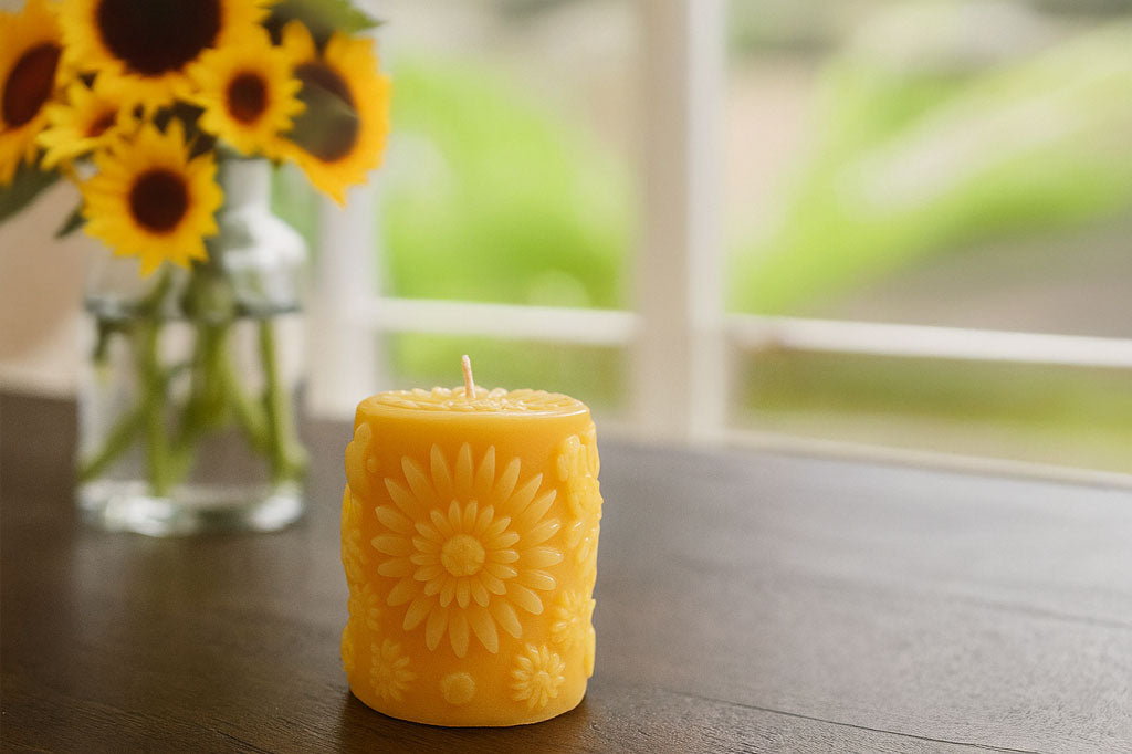Yellow candle with sunflower design on a wooden surface, next to a vase of sunflowers.