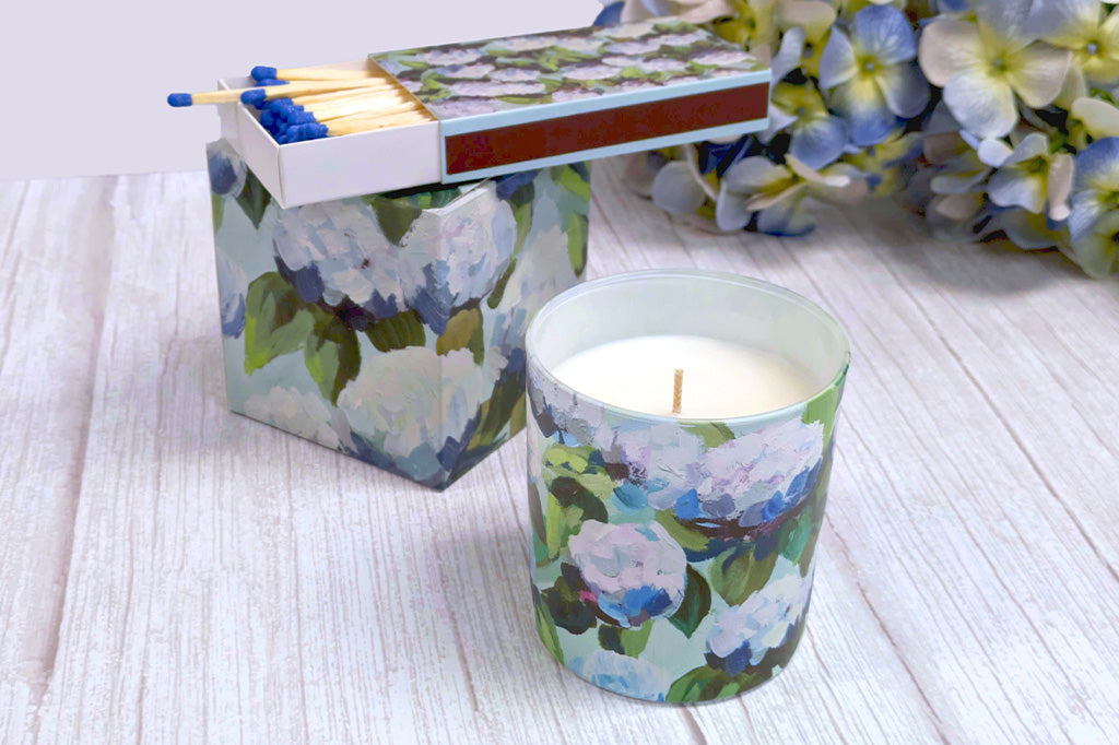 Hydrangea oil painting design on a gift set of colored matches and glass scented candle. Shown on wood table with flowers in background