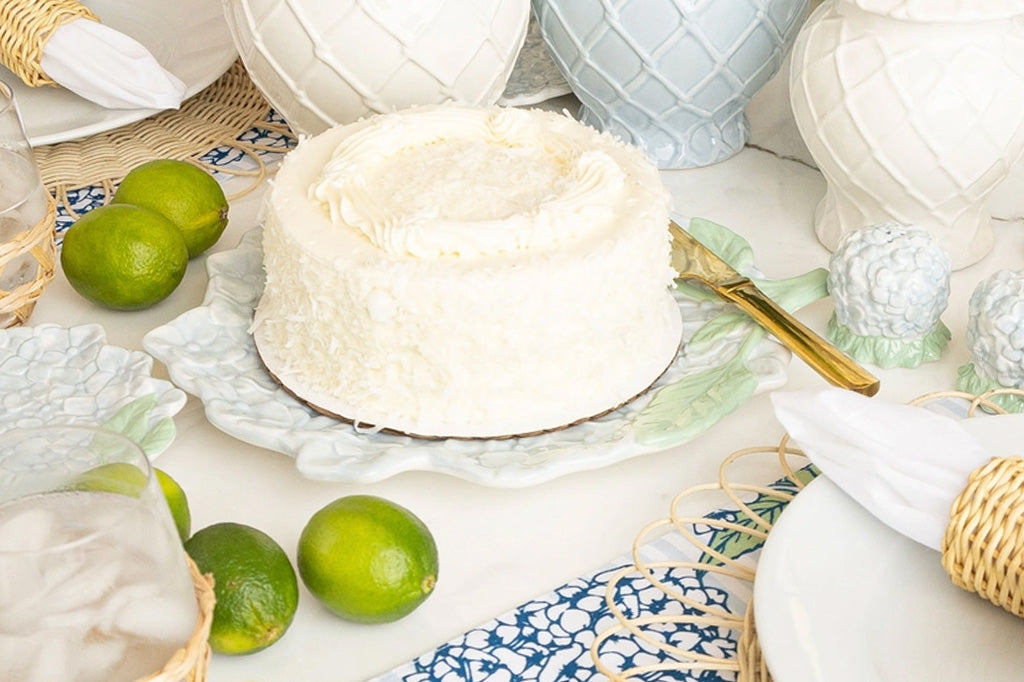 Ceramic serving plate shaped like hydrangea blossom in soft blue with green stem, relief detailing. Shown on festively decorated table with cake on top. Surrounded by wicker place settings and fresh limes in scene.