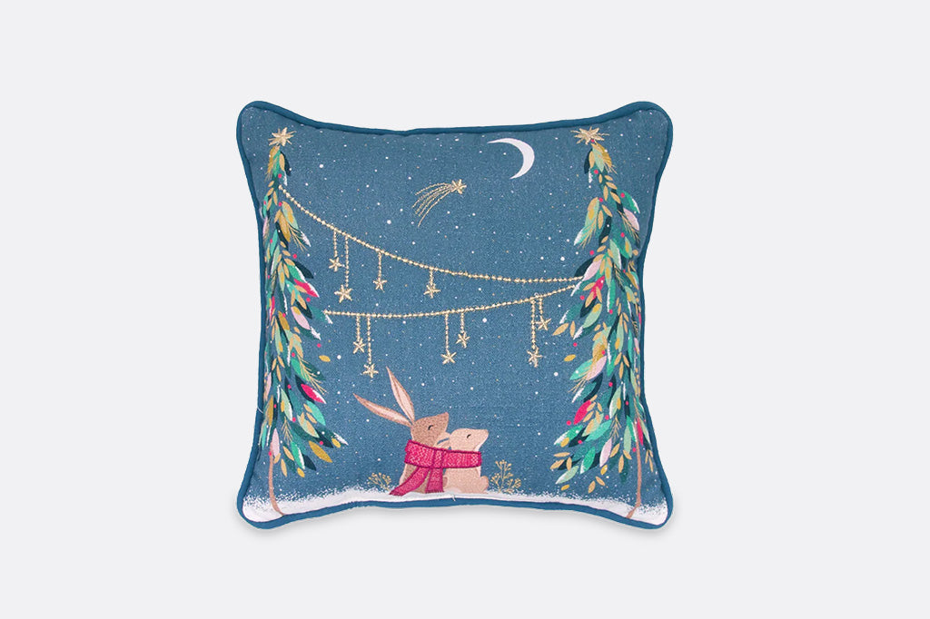 A square decorative throw pillow featuring a winter night scene with a rabbit in a pink scarf under hanging star lights between two colorful trees, on a dark blue background.