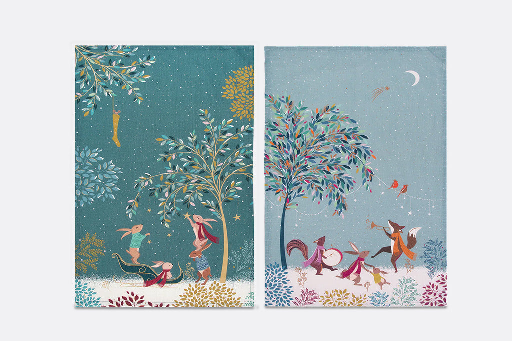 A pair of kitchen towels side by side, featuring whimsical fantasy illustrations. The left towel has a dark teal background with a tree, a fairy, and a small creature in a sleigh.