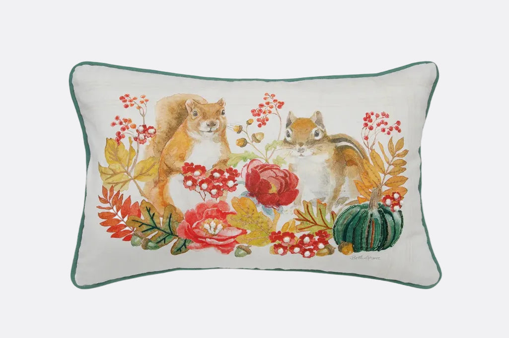 A rectangular decorative throw pillow with a white background and a dark green piped border, featuring an embroidered autumn scene of two squirrels surrounded by red berries, leaves, flowers, and a small green pumpkin.