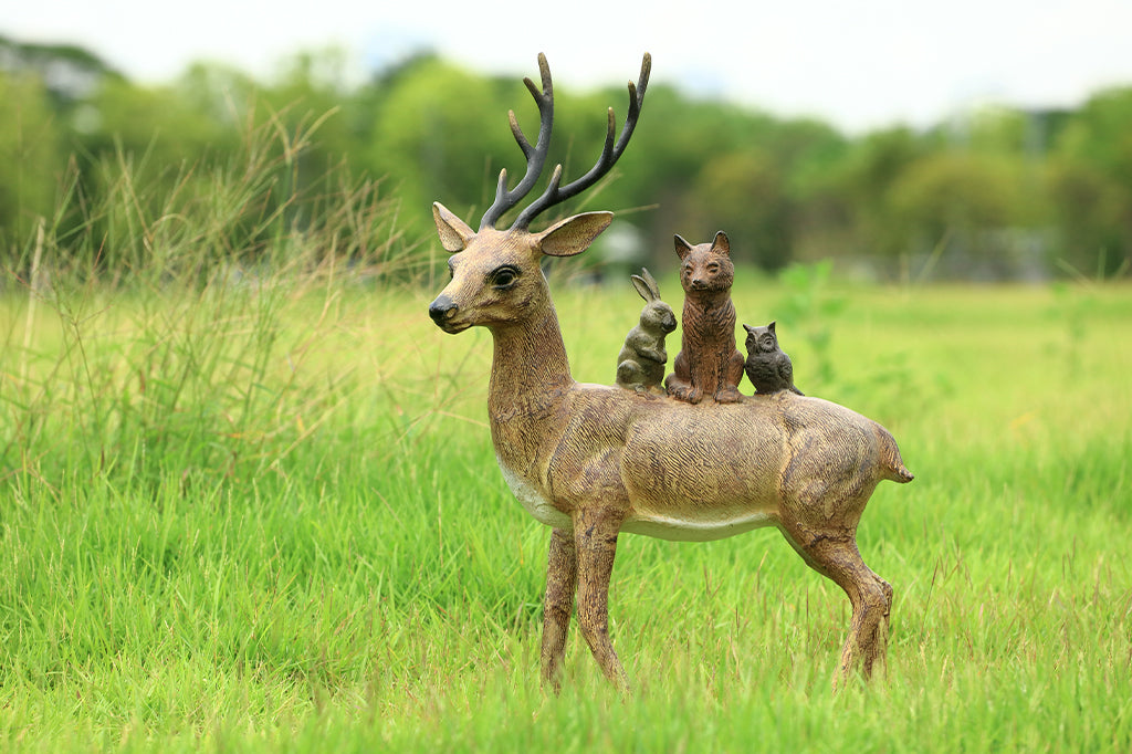 cast metal deer with rabbit, fox, and owl riding on back placed in grassy field. Garden sculpture