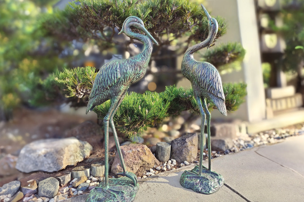 Garden Heron Pair sculpture in front of landscaping and juniper tree