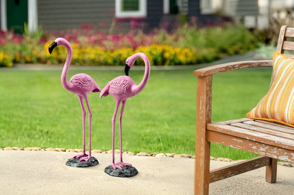 Two pink flamingo statues on a patio with a wooden bench and colorful pillow in the background.