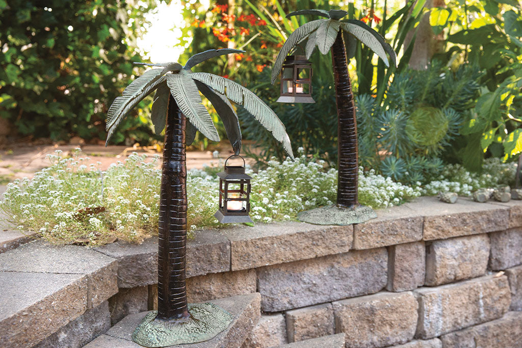 Cast metal palm tree with votive candle lantern hanging from palm leaf, showing a pair of left and right Veradoro Lanterns  in a garden
