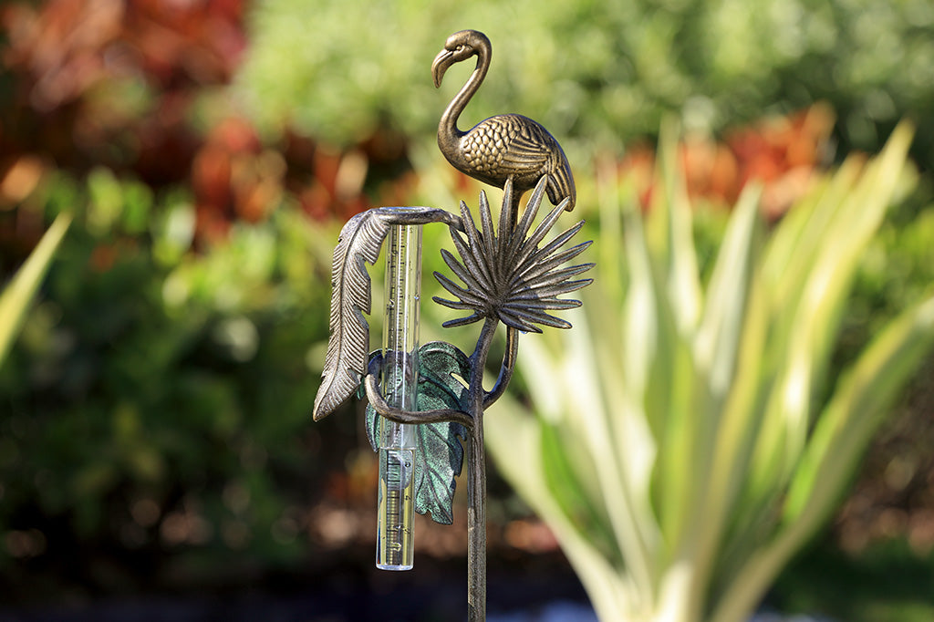 decorative garden rain catcher shows cast metal flamingo, and 3 tropical leaf accents in a garden setting. 