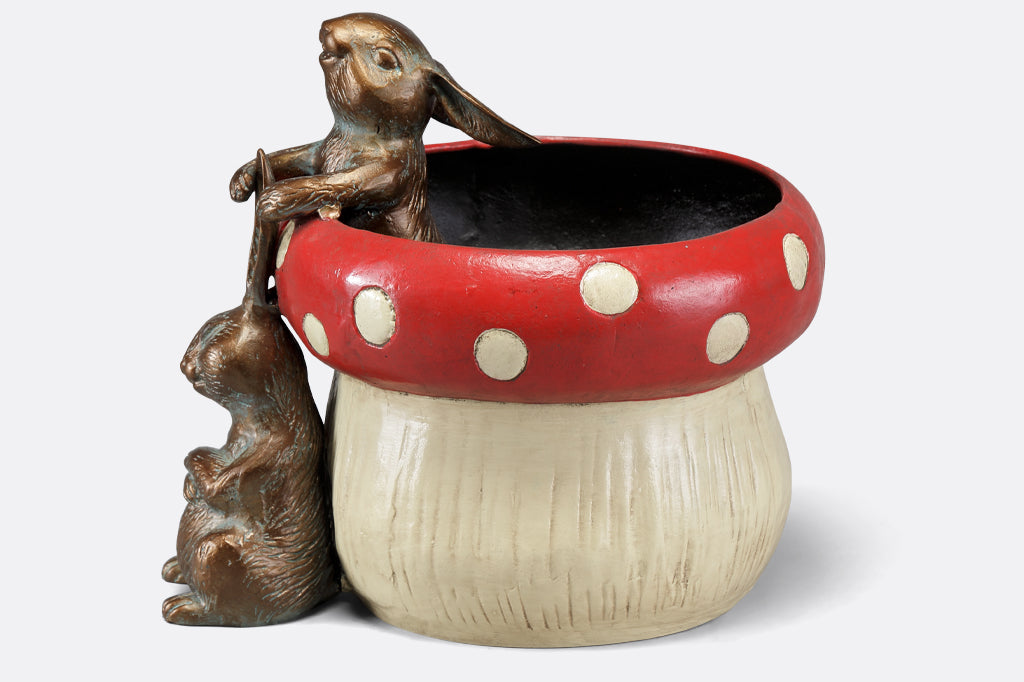 red spotted mushroom planter with two playful bunnies. One bunny peeking outside of planter tugging sitting bunnies ear. Face left