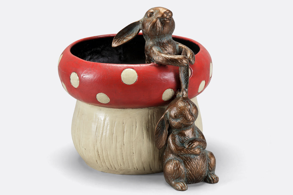 red spotted mushroom planter with two playful bunnies. One bunny peeking outside of planter tugging sitting bunnies ear. 