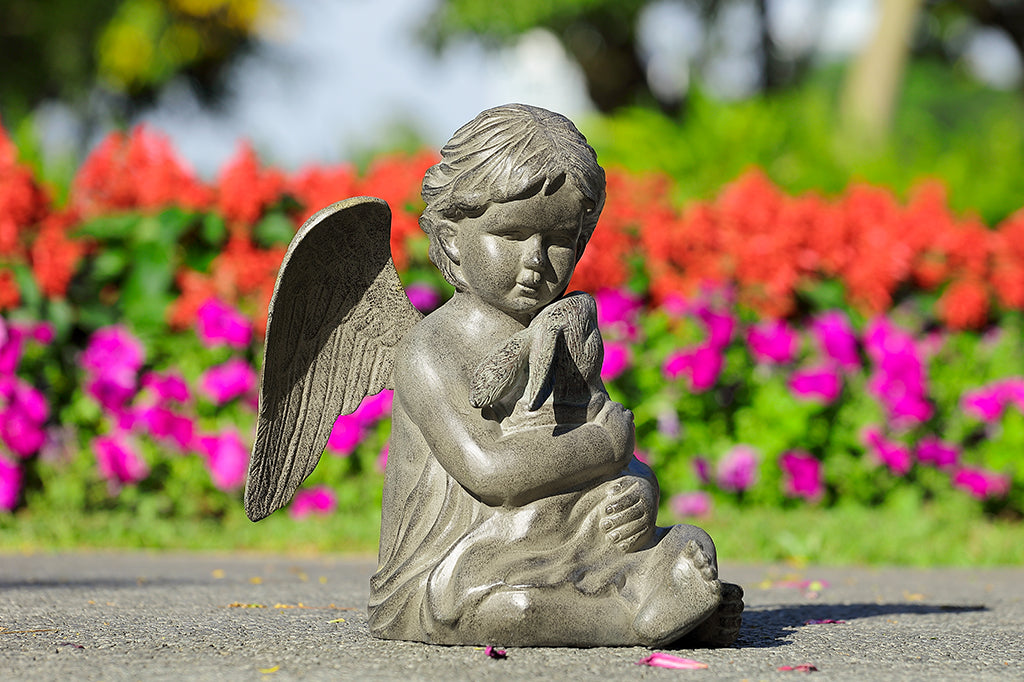 Cast metal cherub holding bunny wrapped in a blanket garden sculpture seated on the pavement against flowers.