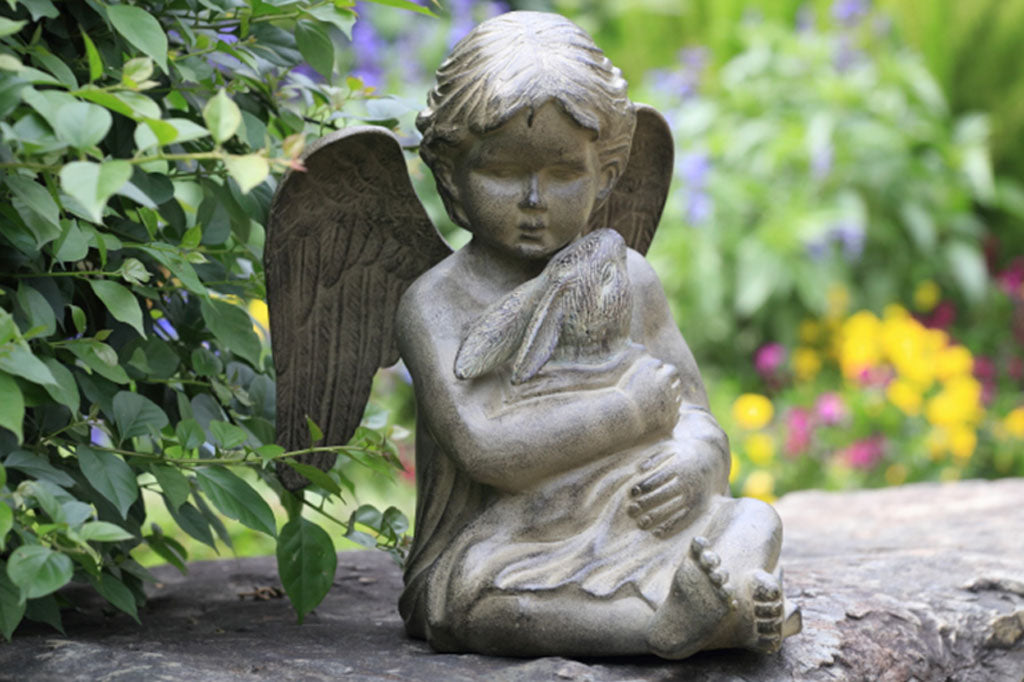 Cast metal cherub baby holding a bunny in a blanket. Set in shade garden on a large stone beside a green shrub