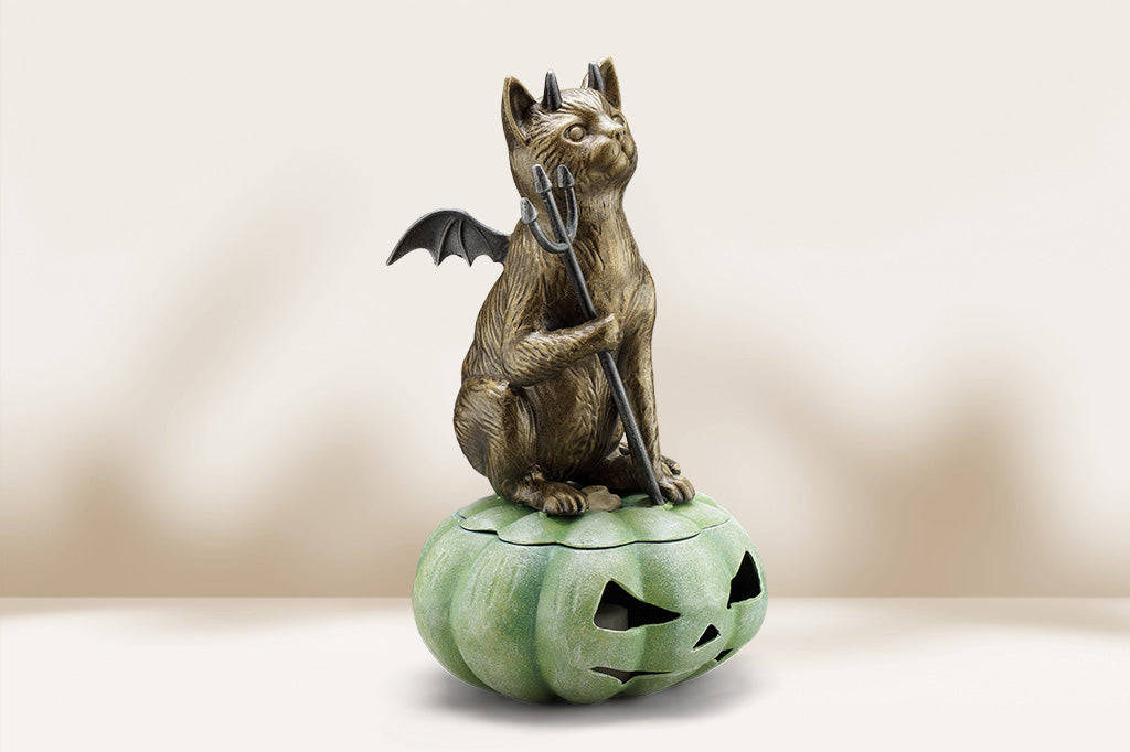 cast metal devil cat with wings sat upon green jack o lantern lantern. It holds a pitchfork.