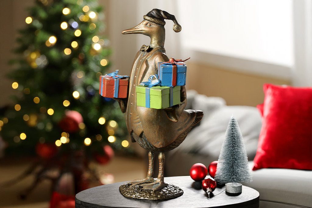 cast metal duck sculpture in formal clothing carrying gift boxes in a living room setting with a tree and lights.